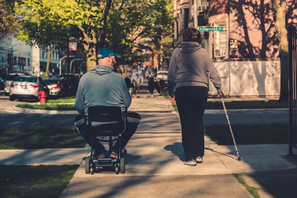 Two people on the sidewalk. One in the wheelchair and one standing.