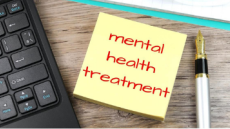 This image shows a corner of a black keyboard on the left hand side, a fountain pen on the right hand side, the head of a clipboard at the top, and in the center a post-it note that reads "Mental Health Treatment" in red ink.