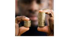 A black man is holding two stacks of gold coins in each hand. The stack of coins in the left hand is smaller than the stack of coins in the right hand. 