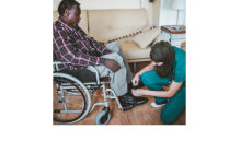 A female direct care worker, who is in scrubs and wearing a hijab, is helping a black man in a wheel chair, who is wearing jeans and a flannel shirt, put on his shoes.