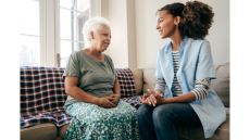 two black women sit on a couch, one older with white hair and one younger. They are smiling at each other