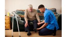 a white older man sits on a couch while a black young man helps him put a shoe on