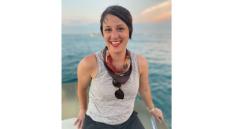 White woman with short brown hair, striped tank top, with a bandana and sunglasses around her neck. she is smiling and is on a boat with water behind her