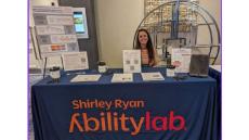 woman seatured behing a conference table with a closth that says shirley ryan abilitylab. the table has papers and pens and the woman has long brown hair and is smiling.