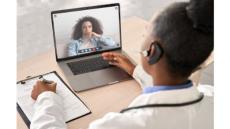 a black female clinician in a white coat looks at a computer where there is a black woman with long hair and a white shirt. they are communicating