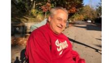 Middle aged man with grey hair wearing a read sweatshirt sitting in a wheelchair outside in Autumn