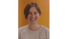 a white woman with short brown hair and a white sweater standing against an orange background. She is smiling.