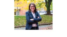 A photo of Carli Friedman, a young white woman with shoulder length brown hair and glasses standing outside