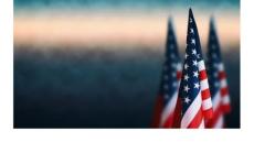 a color photo of the American flag against a blurred background of gray, white and blue sky