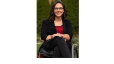 a color photo of Marjorie - a young white woman with glasses and dark brown hair wearing a black suit with a red shirt and sitting in a wheelchair.