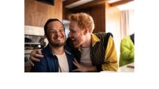 a young man with an intellectual disability is smiling while another young man with red hair holds his shoulders and smiles.