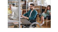 a young man with black hair and a beard is sitting in a wheelchair at a desk typing on a computer