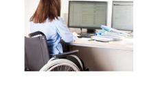 photo of a woman with long brown hair in a wheelchair looking at some computer screens. The photo is from the back so we do not see her face