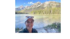color photo of Erin Walaczek, a young white woman waring a white baseball cap with sunglassess on top and a black shirt standing in front of a lake