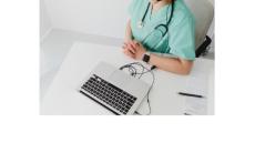 Photo of a person wearing blue scrubs sitting at a table in front of a laptop. Their head isn't shown in the picture. The person is white and has a stethoscope around their neck and a smart watch