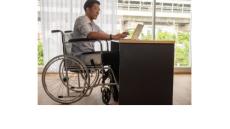 A photo of am Asian man in a wheelchair at a desk working on his laptop