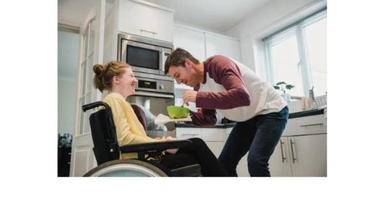 A young woman with red hair in a bun wearing a yellow sweatshrit sits in a wheelchair as a young man in jeans and a red and white shirt hold a green bowl in front of her.