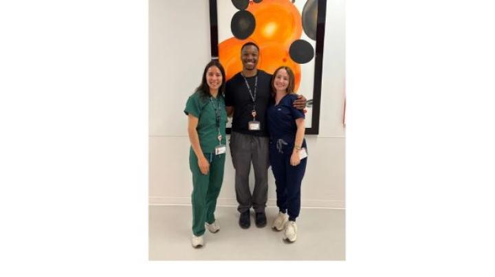 group photo of medical externs - a young white woman in green scrubs with dark hair is on the left, a youhg Black man in gray pants and a black top is in the middle, and a young white woman in navy scrubs with brown hair is on the right.