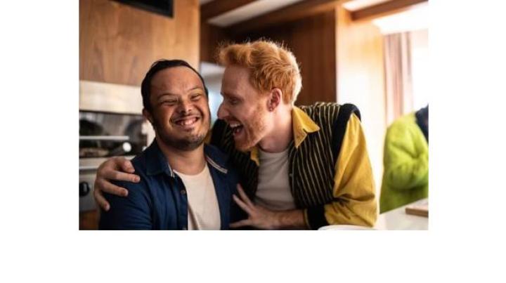 a young man with an intellectual disability is smiling while another young man with red hair holds his shoulders and smiles.