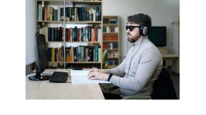 a white man with sunglassess and headphones sitting at a computer in a room with bookshelves in the background