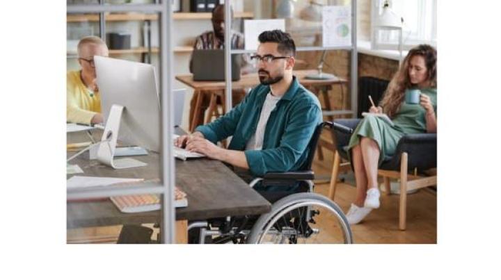 a young man with black hair and a beard is sitting in a wheelchair at a desk typing on a computer
