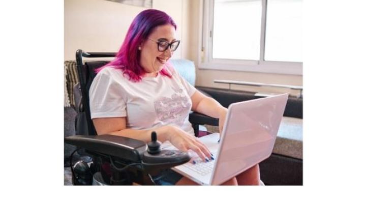 color photo of a white woman with pink hair, black glasses and a white top sitting in a wheelchair working at a computer.