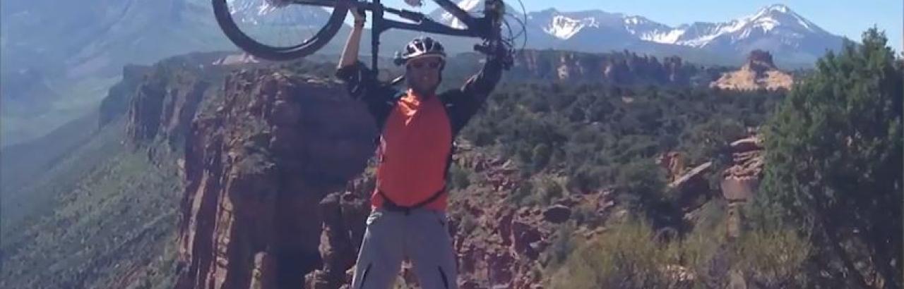 Sami on a mountain joyously holding a bike above his head