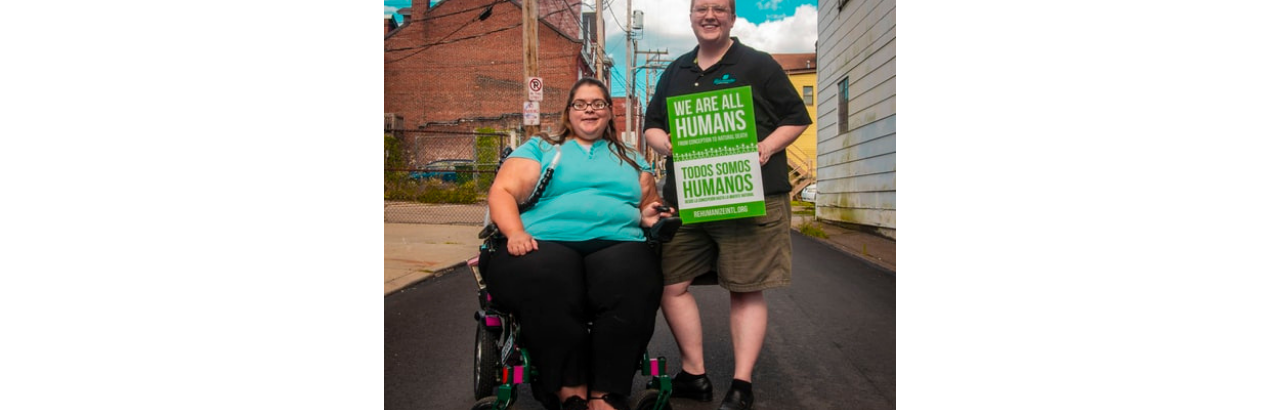 A person with a disability is sitting in a wheelchair while standing next to someone holding a green sign that reads "We are all human" in English and Spanish. 