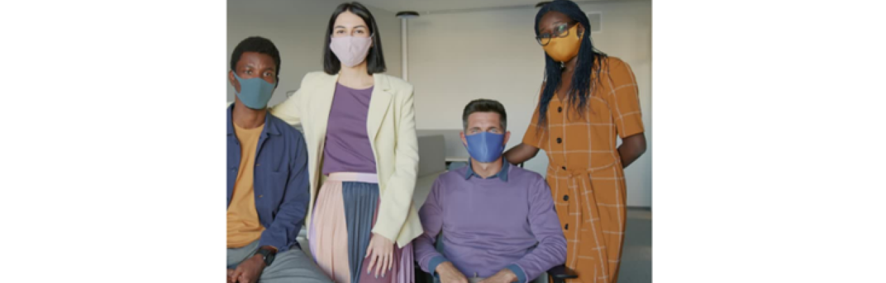A young black man is sitting to the left of a standing white female, who is standing next to a white man sitting in a wheelchair, who is next to a standing young black woman. All of the individuals are looking into the camera and have a face mask on. 