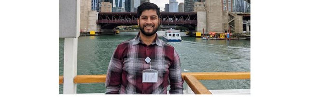 This is a picture of Dhrumil Shah. He is an Indian man who is wearing a Red flannel shirt. Behind him are a series of skyscrapers in Chicago.