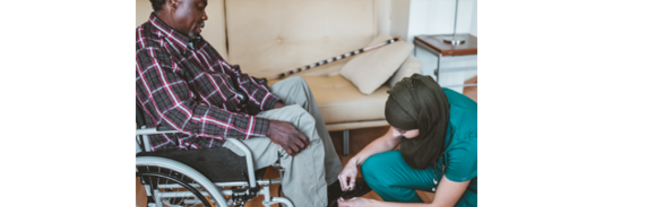 A female direct care worker, who is in scrubs and wearing a hijab, is helping a black man in a wheel chair, who is wearing jeans and a flannel shirt, put on his shoes.