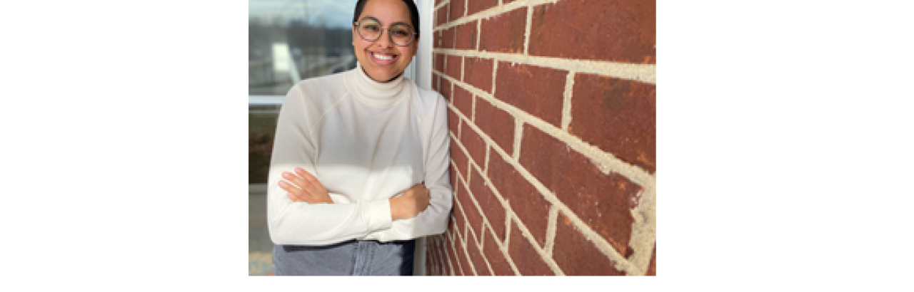 Niveda - young woman in white turtleneck smiling and leaning against a brick wall