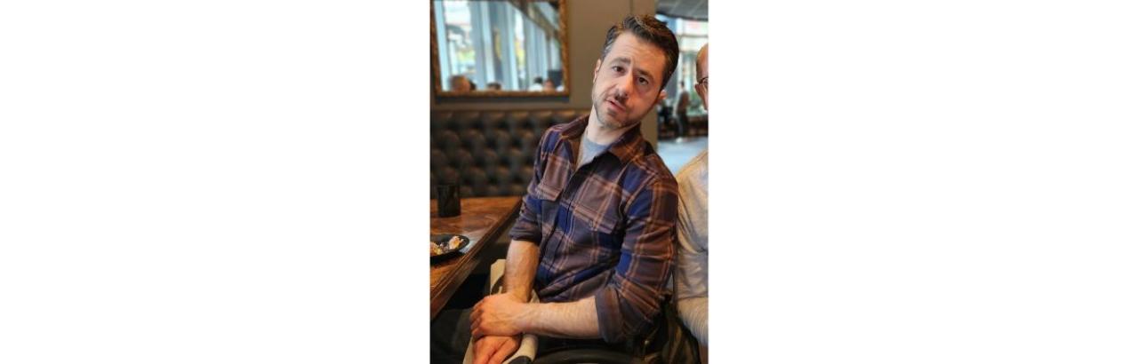 Man with brown hair and beard sitting in a restaurant wearing a plaid shirt
