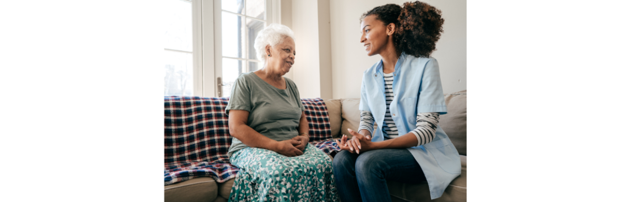 two black women sit on a couch, one older with white hair and one younger. They are smiling at each other