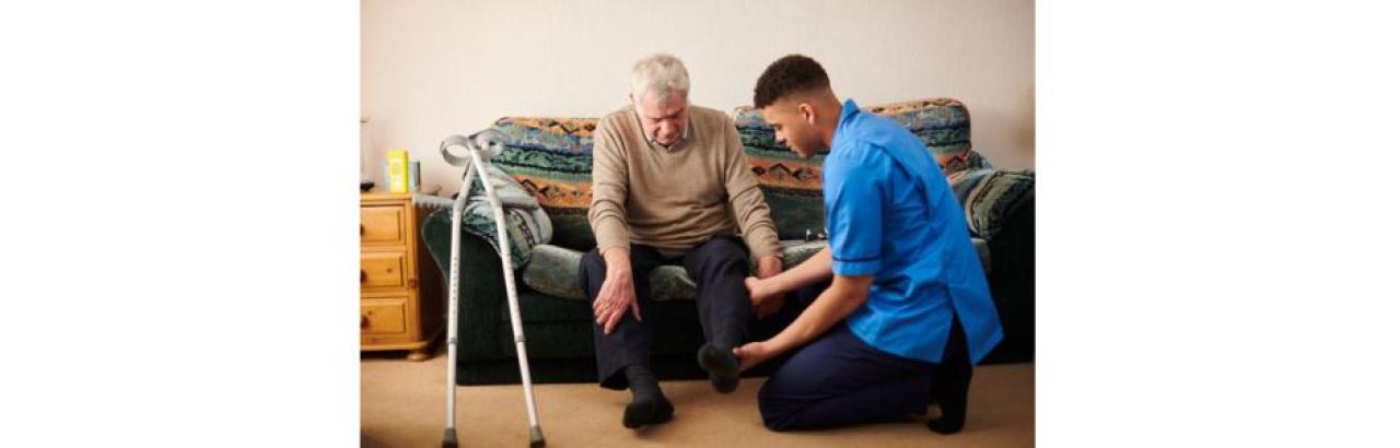 a white older man sits on a couch while a black young man helps him put a shoe on