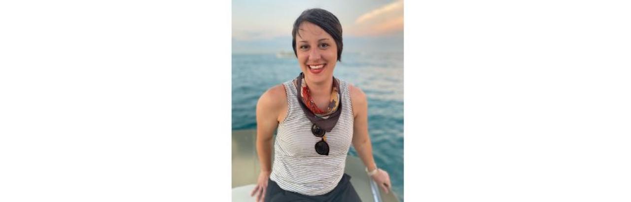 White woman with short brown hair, striped tank top, with a bandana and sunglasses around her neck. she is smiling and is on a boat with water behind her