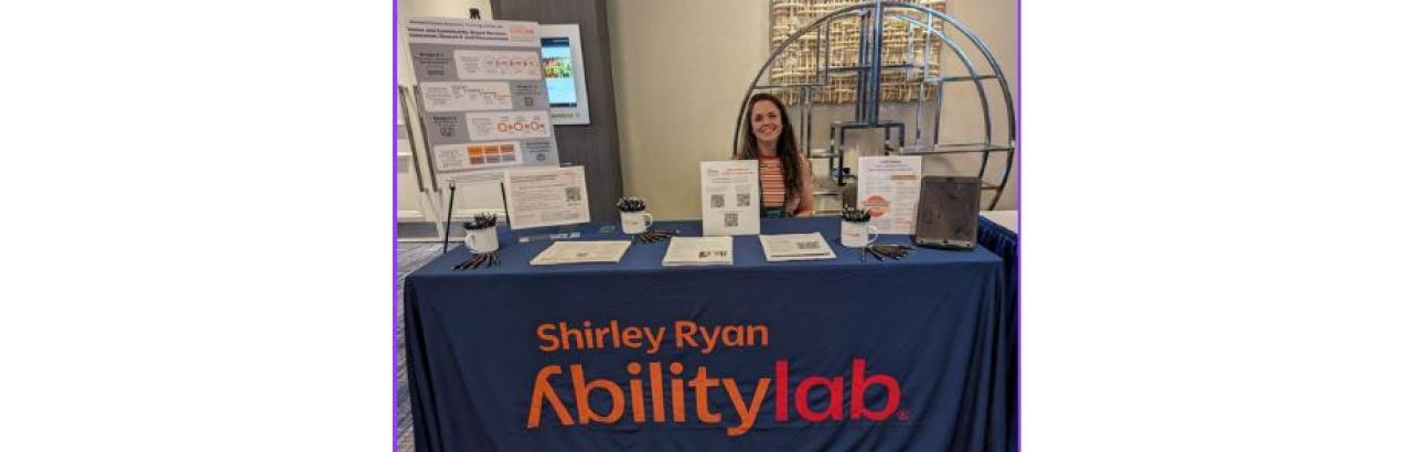 woman seatured behing a conference table with a closth that says shirley ryan abilitylab. the table has papers and pens and the woman has long brown hair and is smiling.