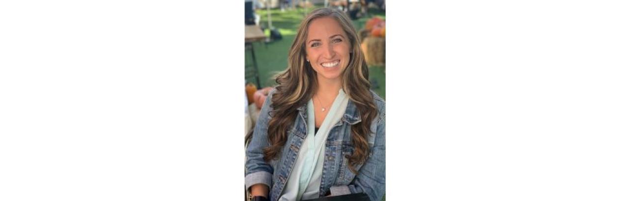 young white woman with long brown hair wearing a jean jacket and white shirt underneath is smiling. She is in an outdoor setting.