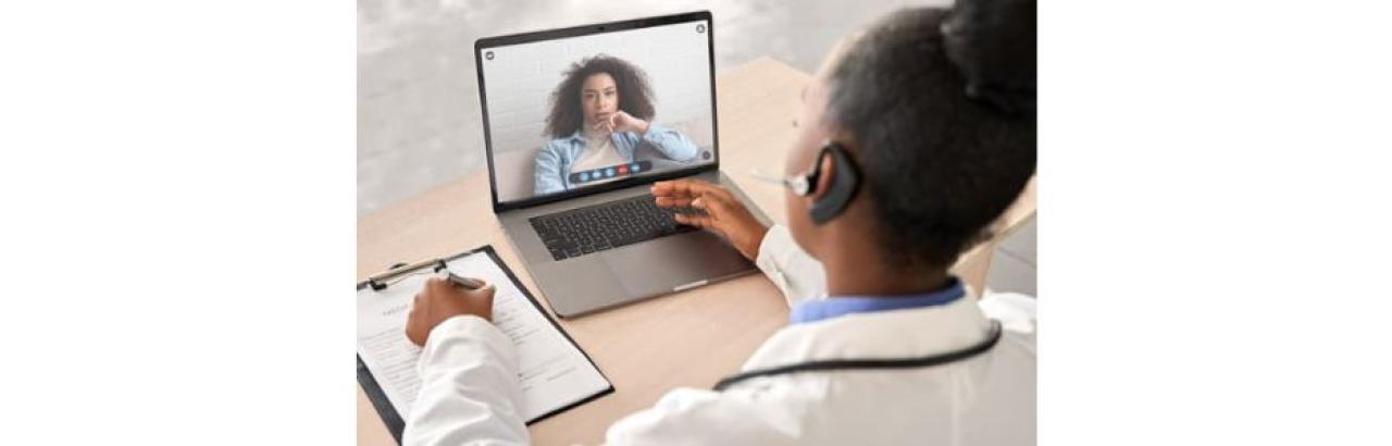 a black female clinician in a white coat looks at a computer where there is a black woman with long hair and a white shirt. they are communicating