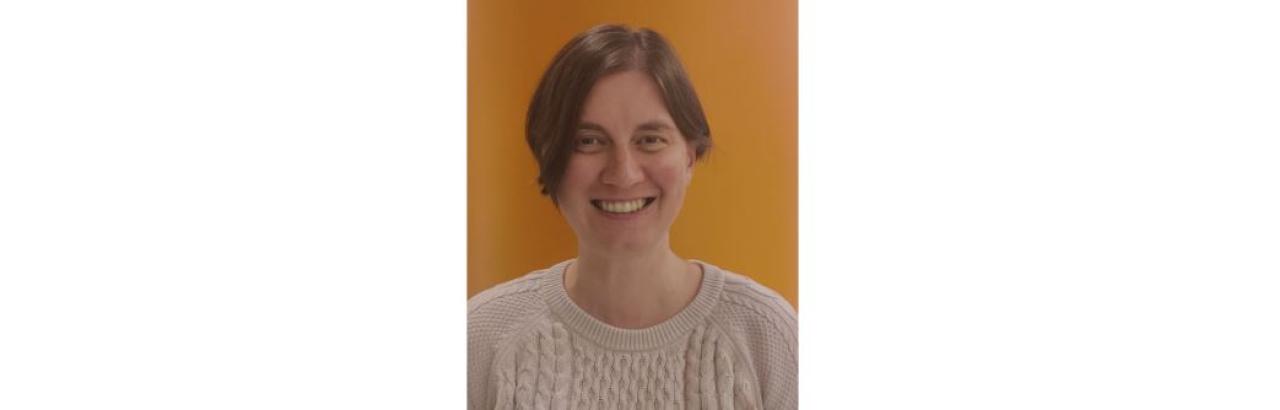 a white woman with short brown hair and a white sweater standing against an orange background. She is smiling.