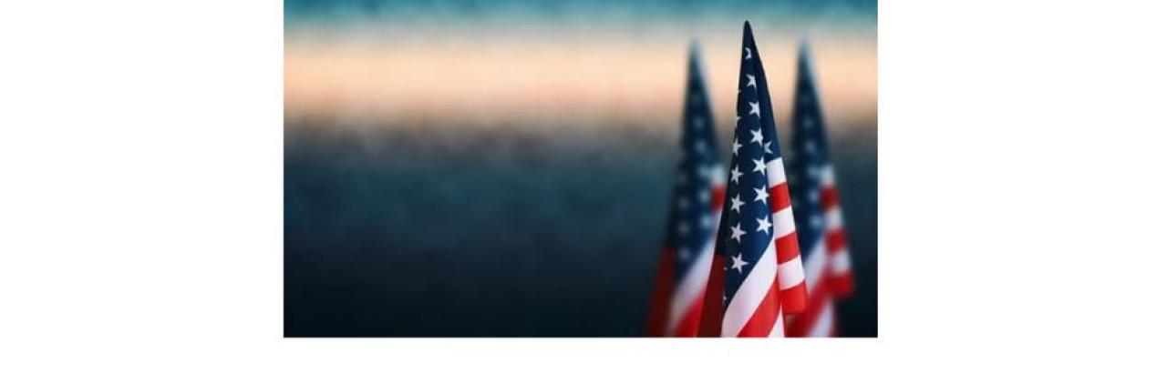 a color photo of the American flag against a blurred background of gray, white and blue sky