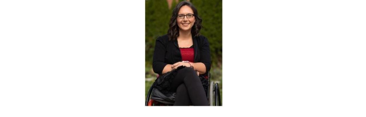 a color photo of Marjorie - a young white woman with glasses and dark brown hair wearing a black suit with a red shirt and sitting in a wheelchair.