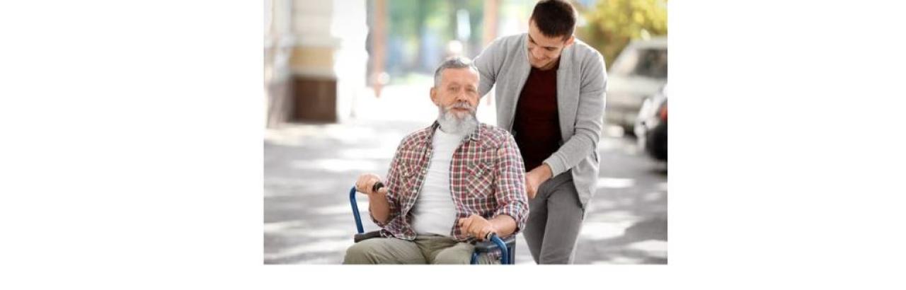 photo of an older white man in a wheelchair with a younger white man leaning over him to talk