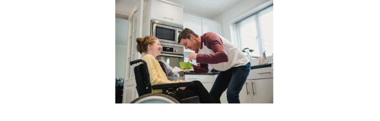 A young woman with red hair in a bun wearing a yellow sweatshrit sits in a wheelchair as a young man in jeans and a red and white shirt hold a green bowl in front of her.