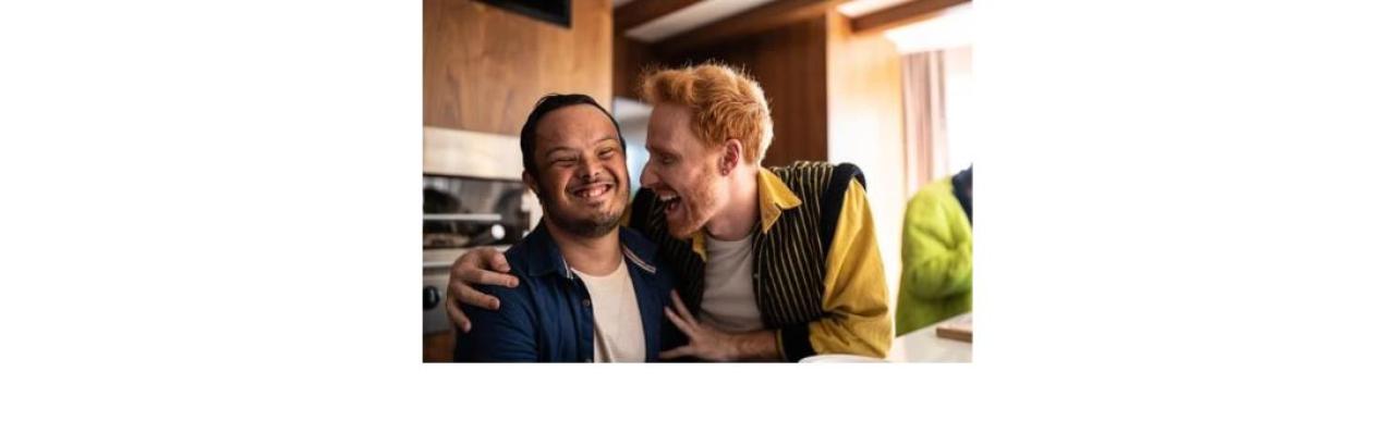 a young man with an intellectual disability is smiling while another young man with red hair holds his shoulders and smiles.