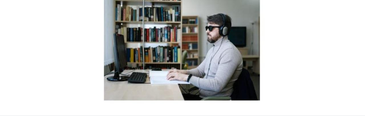 a white man with sunglassess and headphones sitting at a computer in a room with bookshelves in the background