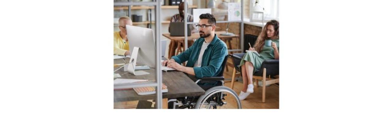 a young man with black hair and a beard is sitting in a wheelchair at a desk typing on a computer