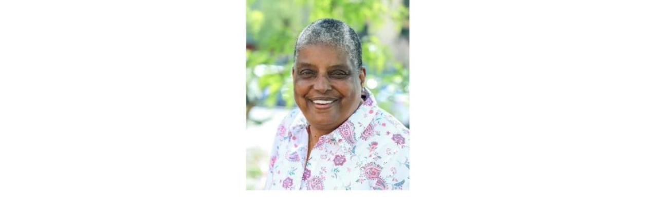 color photograph of Cindy Bently - a middle aged Black woman with short grey hair wearing a white top. She is outside and is smiling