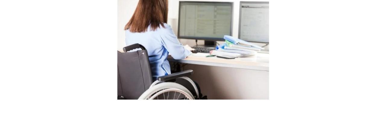 photo of a woman with long brown hair in a wheelchair looking at some computer screens. The photo is from the back so we do not see her face