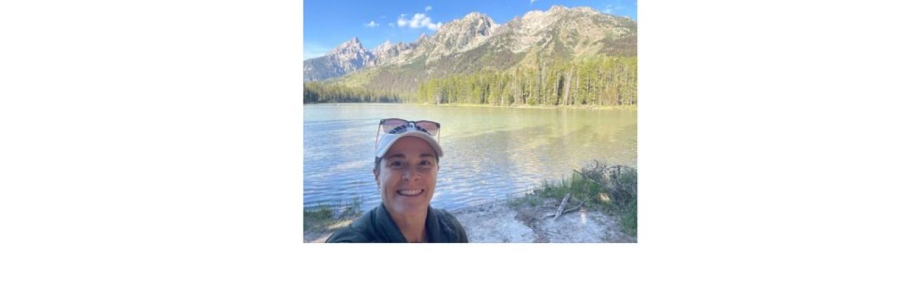 color photo of Erin Walaczek, a young white woman waring a white baseball cap with sunglassess on top and a black shirt standing in front of a lake